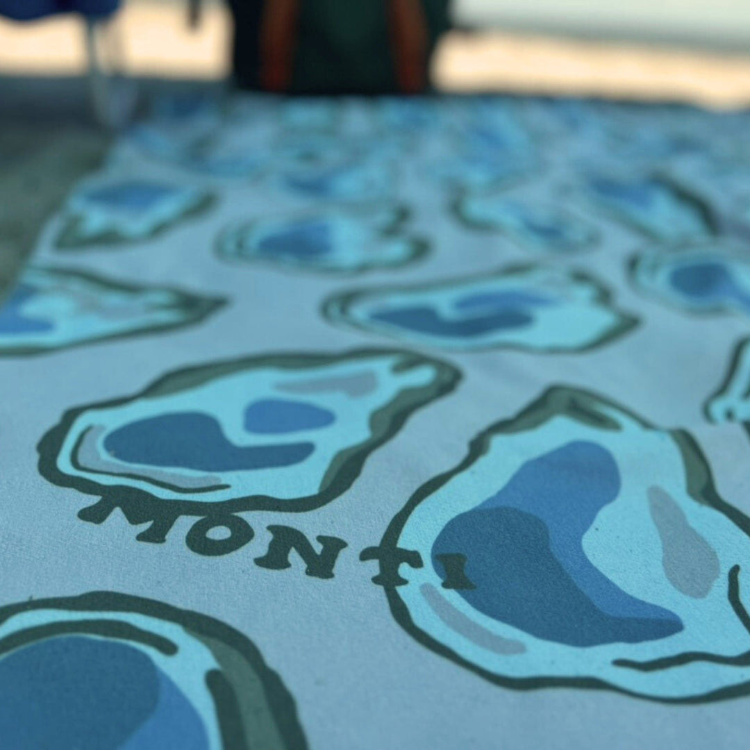 Monti Coolers Big Ass Towel in oyster color, large absorbent beach towel with soft fabric and bold branding, laid out on sand.